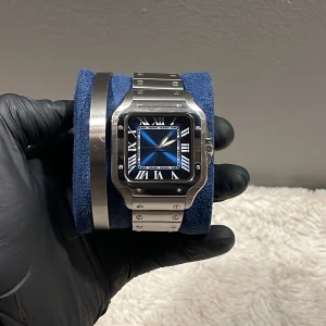 Silverfärgad klocka med blå urtavla - Elegant klocka med en fyrkantig boett i silver och en stilren blå urtavla. Klockan har romerska siffror och ett matchande silverfärgat armband. Perfekt för den som vill ha en tidlös accessoar.