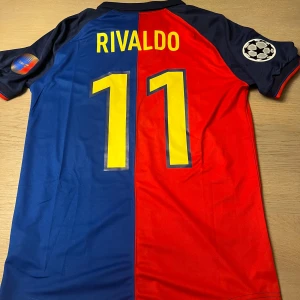 Blå och röd fotbollströja från Nike - Säljer en klassisk fotbollströja från Nike i blått och rött med gula detaljer. Tröjan har korta ärmar och ett tryck med 'Rivaldo 11' på ryggen. Framsidan har klubbmärke och årtalen 1899-1999. Perfekt för fotbollsfans!
