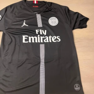 Svart PSG fotbollströja - Snygg svart PSG fotbollströja med Neymar Jr och nummer 10 på ryggen. Tröjan har korta ärmar och Fly Emirates som sponsorlogga på framsidan. Perfekt för fotbollsfans!