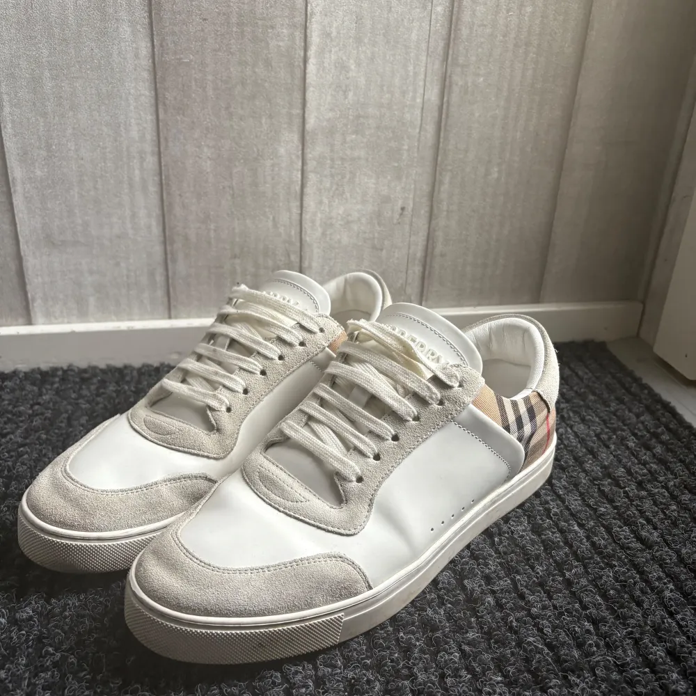 Snygga vita sneakers från Burberry med klassiskt rutigt mönster på hälen. Skorna har snörning och en stilren design med detaljer i mocka och syntetmaterial. Perfekta för en trendig look. Köpte för 6800 Allt Og ingår. Kengät.