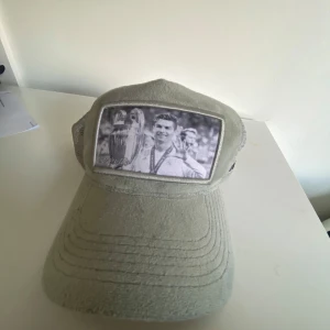 Grå keps med fototryck - Snygg grå keps med ett svartvitt fototryck på Cristiano Ronaldo. Kepsen har ett nätmaterial på baksidan för bättre ventilation och justerbar passform. 9/10 Skick Hör av dig vid fler funderingar