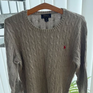 Grå stickad tröja från Ralph Lauren - Säljer en klassisk beige stickad tröja från Ralph Lauren med kabelstickat mönster. Tröjan har långa ärmar och en rund halsringning. Perfekt för kyligare dagar. 🧶