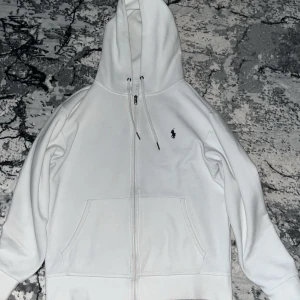 Vit hoodie från Ralph Lauren - Säljer en stilren vit hoodie från Ralph Lauren med dragkedja och klassisk logga på bröstet. Den har en bekväm passform och praktiska fickor framtill. Perfekt för en avslappnad look.