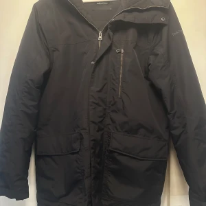 Svart parkajacka från Peak Performance - Säljer en stilren svart vinterjacka från Peak Performance. Jackan har en praktisk dragkedja och flera fickor, inklusive en bröstficka med dragkedja. Perfekt för kyligare dagar med sin långa ärm och skyddande huva. 