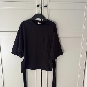 Svart topp från Nelly - Säljer en svart topp från Nelly med korta ärmar och knytband i ryggen💖 Perfekt för sommaren. Toppen har en rund halsringning och är tillverkad i ett mjukt material. Endast använd 1 gång🩷