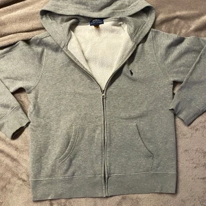 Grå hoodie från Polo Ralph Lauren - Säljer en stilren grå hoodie från Polo Ralph Lauren med dragkedja och broderad logga på bröstet. Tröjan har en mjuk insida och långa ärmar, perfekt för en avslappnad stil. 🐎Ungdom Str 14-16.