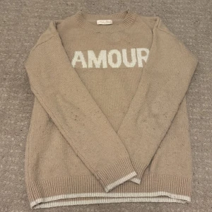 Beige stickad tröja från Kids Star - Säljer en beige stickad tröja från Kids Star med texten 'AMOUR' i vitt på framsidan. Tröjan har långa ärmar och ribbstickade muddar vid ärmslut och nederkant. Perfekt för en mysig dag! Har ett super litet hål som är insytt och syns inte alls😍