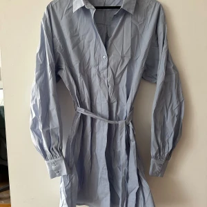 Ljusblå skjortklänning från Rut&Circle - Snygg ljusblå skjortklänning från Rut&Circle med knappar framtill och ett matchande bälte i midjan. Klänningen har långa ärmar med knappar vid manschetterna och en klassisk krage. Perfekt för en stilren look! Lite skrynklig på bilderna pga den legat i en låda på vinden och jag ej strukit den. Den är jättefin när den väl är struken!
