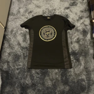 Svart t-shirt från Glorious Gangsta - Snygg svart t-shirt från Glorious Gangsta med ett stort, guldigt emblem på bröstet. T-shirten har korta ärmar och en normal passform. Perfekt för en stilren look. och den är xs men passar som S.