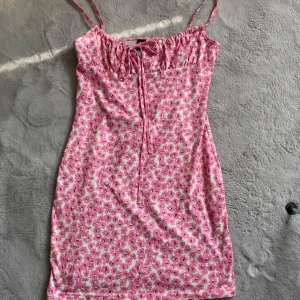 Rosa blommig klänning från Shein - Söt rosa klänning från Shein med blommigt mönster och smala axelband. Klänningen har en knytning framtill och är perfekt för varma dagar. Den är tillverkad i ett lätt och bekvämt material.