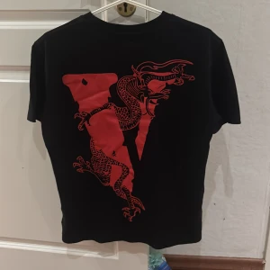 Svart t-shirt med röd drake och VLONE-tryck - Säljer en svart t-shirt från VLONE med en stor röd drake på baksidan och ett stort VLONE-tryck på framsidan. T-shirten har korta ärmar och är perfekt för en cool och avslappnad stil.