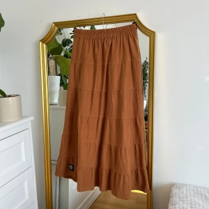 Brun/orange långkjol  - Säljer en snygg brun långkjol från Stockholm. Kjolen har en elastisk midja och flera volangdetaljer som ger en fin struktur. Perfekt för en avslappnad stil. Passar bra till både enkla toppar och mer dressade överdelar. Står ingen storlek i kjolen och har ett minna av att den är ”one size” Jag skulle säga att den passar dem från XS-M