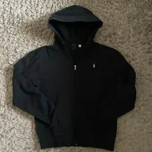 Svart hoodie från Ralph Lauren - Säljer en stilren svart hoodie från Ralph Lauren med dragkedja och vit logga. Perfekt för en avslappnad look. Storlek M. Nypris ca 2300kr