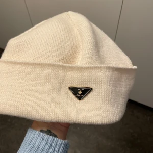 Vit mössa från Canada Snow - Snygg vit mössa från Canada Snow med en stilren design. Mössan har en liten svart och guld triangel-logga framtill. Perfekt för kyliga dagar.