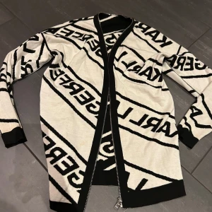 Svartvit kofta från Karl Lagerfeld - Snygg svartvit kofta från Karl Lagerfeld med ett iögonfallande mönster av märkesnamnet. Koftan har en dragkedja framtill och långa ärmar. Perfekt för att ge din outfit en stilren touch.