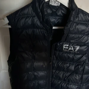 Fet Merin blå dunväst från EA7 - Snygg merinblå dunväst från Emporio Armani med dragkedja och quiltad design. Perfekt för varmare dagar och ger en stilren look. Västen har en praktisk förvaringspåse med logotyp.