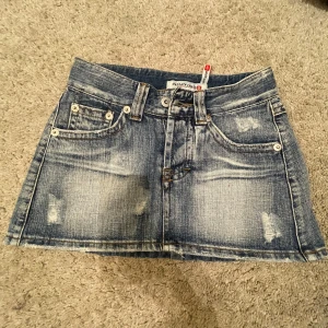 Blå jeanskjol från Gassata - Snygg blå jeanskjol från Gassata med slitna detaljer och klassisk femficksdesign. Kjolen har en kort längd och stängs med knapp och dragkedja framtill. Perfekt för en avslappnad stil!