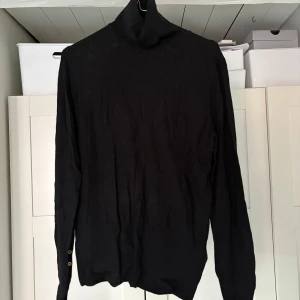 Svart polotröja med knappar - Säljer en stilren svart tunn polotröja med långa ärmar och dekorativa guldknappar vid ärmsluten. Perfekt för en elegant look. Tröjan är i ett mjukt material som ger en bekväm passform.