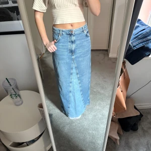Lång jeanskjol från Zara - Säljer en snygg lång jeanskjol från Zara i klassisk blå denim. Kjolen har en rak passform med femficksdesign och knappgylf. Perfekt för en avslappnad stil.