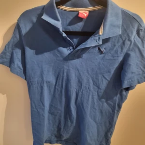 Blå pikétröja från Puma stl 164 - Blå pikétröja från Puma med klassisk krage och knappar. Perfekt för en avslappnad stil. Tröjan har korta ärmar och en liten Puma-logga på bröstet.