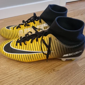 Gula fotbollsskor från Nike - Gula Nike Mercurial fotbollsskor med svarta och vita detaljer. Skorna har snörning och en hög ankelkrage för extra stöd. Till gräs. Stl Eur 43 UK, 8,5 US 9,5