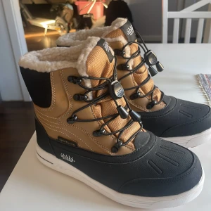Vinterkängor från Áhkká - Snygga och varma vinterkängor från Áhkká i svart och beige med fuskpälsfoder. De har snörning och en robust sula för bra grepp. Perfekta för kalla dagar!