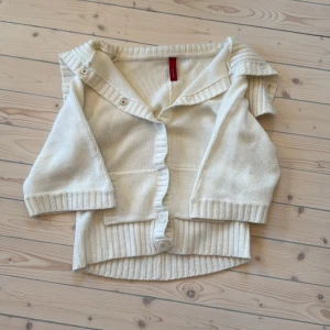 Vit kofta med knappar - Säljer en vit kofta med knappar framtill och en bred krage som kan vikas ner. Koftan har trekvartsärmar och ribbade detaljer vid ärmslut och nederkant. Perfekt för en stilren look.