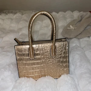 Guldig handväska - Snygg guldig handväska med krokodilpräglat mönster och dragkedja. Perfekt för att ge en lyxig touch till din outfit. Väskan har två handtag och en praktisk storlek för att bära dina viktigaste saker💓