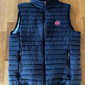 Mörkblå dunväst från Colmar - Snygg mörkblå dunväst från Colmar med horisontella sömmar och dragkedja framtill. Västen har två fickor med dragkedja och en liten Colmar-logga på bröstet. Insidan är ljusgrå och västen är lätt och smidig att bära.