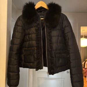 Svart dunjacka med pälsdetaljer - Snygg svart dunjacka med fluffig päls runt kragen och dragkedja framtill. Jackan har långa ärmar och quiltad design med dekorativa fransar på fickorna. Perfekt för kyliga dagar.