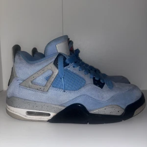 University Blue Nike Air Jordan 4s - Snygga Nike Air Jordans 4s i ljusblå mocka med grå och svarta detaljer. Skorna har en klassisk snörning och den ikoniska Jumpman-loggan på hälen. Perfekta för dig som vill ha en stilren och sportig look.