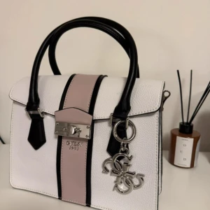 Vit och rosa handväska från Guess - Använd endast 4 gånger  Elegant handväska från Guess med en stilren design i vitt med svarta detaljer på handtagen och kanterna. Väskan har en dekorativ rem i en ljusrosa nyans och en silverfärgad Guess-logotyp framtill. En charmig berlock med Guess-loggan hänger från handtaget, vilket ger en extra touch av lyx. Väskan har både handtag och en avtagbar axelrem för olika bäralternativ. Insidan är rymlig med flera fack för att organisera dina tillhörigheter.
