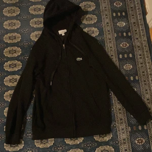 Svart hoodie från Lacoste - Säljer en stilren svart hoodie från Lacoste med dragkedja och huva. Den har det klassiska Lacoste-logotypen på bröstet. Perfekt för en avslappnad stil.
