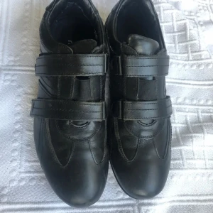 Svarta skinn sneakers med kardborreband - Snygga svarta sneakers med praktiska kardborreband för enkel på- och avtagning. Skorna har en stilren design med sömdetaljer och en slitstark sula. Perfekta för en casual look.