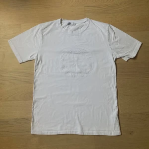 Vit Stone Island t-shirt med relieftryck - Vit t-shirt från Stone Island med stort, diskret relieftryck av loggan på bröstet. Klassisk rund hals och korta ärmar. Tillverkad i mjuk bomull som känns skön mot huden. Perfekt för dig som gillar clean och stilren streetwear.