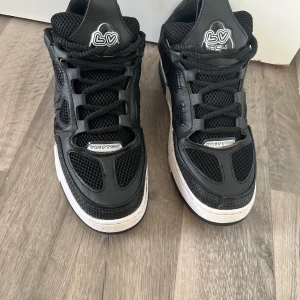 Louis Vuitton svarta sneakers 9.5 - Louis Vuitton sneakers i svart med vita detaljer och coola LV-loggor på tungan och hälen. Skorna har meshpaneler, snörning och en platt sula med mönstrade sidopaneler. Tillverkade i Italien med en mix av skinn och syntetmaterial. Perfekt för dig som gillar exklusiv streetstyle.