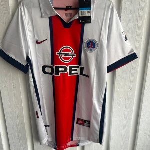 PSG Okocha 10 retro fotbollströja Nike - Snygg PSG fotbollströja med Okocha 10 på ryggen, tillverkad av Nike. Tröjan har vit bas med bred röd mittremsa och mörkblå detaljer, samt klubbmärke och Opel-logga på bröstet. Kortärmad modell i lätt och glansigt material, perfekt för dig som gillar klassiska retrotröjor.