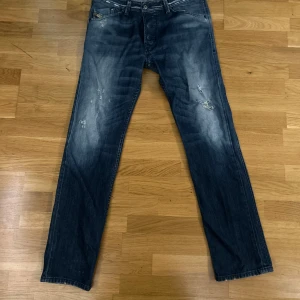 Blå jeans från Diesel - Säljer ett par snygga och unika blå jeans från Diesel i modellen Darron. De har en cool, sliten look med ljusa tvättade detaljer och klassiska fem fickor. Perfekta för en avslappnad stil, oavsett om du ska på stan eller hänga med kompisar. Passformen är normal och de är tillverkade i slitstarkt denim.
