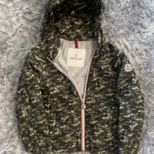 Moncler camo vindjacka med huva - Snygg vindjacka från Moncler i camouflagemönster med gröna och beige toner. Jackan har huva, hel dragkedja med Moncler-logga och ett grått innerfoder. Patch med Moncler-emblem på ärmen ger extra streetkänsla. Perfekt för dig som gillar att sticka ut.