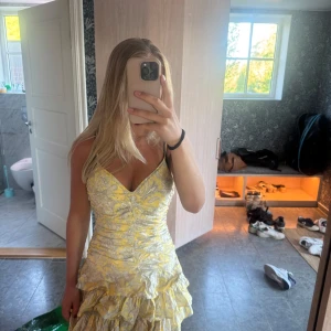 Gul och vit kortklänning med volanger - Supersöt kortklänning i gul och vit med blommigt mönster och volanger nertill. Klänningen har v-ringad urringning och smala axelband, samt en figurnära passform som framhäver midjan. Perfekt för dig som gillar färg och detaljer!