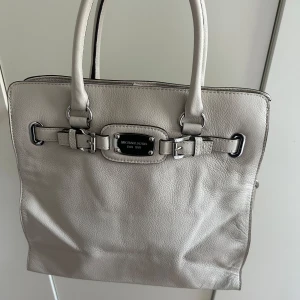 Ljusgrå väska från Michael Kors - Snygg ljusgrå väska från Michael Kors i mjukt skinn med silverfärgade metalldetaljer och dekorativt spänne framtill. Väskan har både handtag och axelrem, samt flera fack och dragkedja inuti för smart organisering. Perfekt storlek för allt du behöver