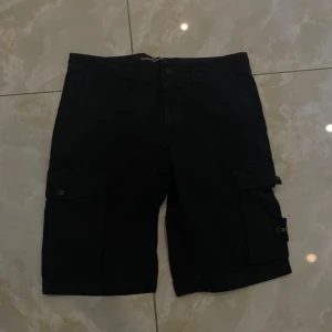 Svarta cargo shorts från Stone Island - Svarta cargo shorts från Stone Island med klassisk logopatch på benet. De har flera fickor, inklusive stora sidofickor med knappstängning. Shortsens material är bomull och de har en normal passform med midwaist. Perfekta för chill dagar och streetwear.