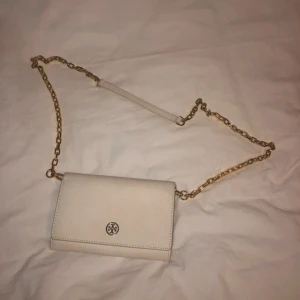 Vit axelväska från Tory Burch - Snygg beige axelväska från Tory Burch med guldfärgad kedja och klassisk logga framtill. Väskan är i slätt skinn med stilren design och har ett praktiskt axelband med kedjedetaljer. TYVÄRR HAR VÄSKAN SMÅ DEFEKTER. De två sista bilderna visar defekterna
