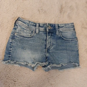 Blå jeansshorts med slitningar  - Snygga blå low waist jeansshorts med fransig kant och lite slitningar. På ena bakfickan finns en fjäril. Sortsen har knapp och dragkedja framtill samt bälteshällor. De är från h&m i storlek 134/140.