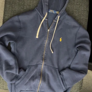 Blå hoodie från Polo Ralph Lauren - Snygg marinblå hoodie från Polo Ralph Lauren med gul broderad logga på bröstet. Tröjan har huva med vita snören, dragkedja framtill och två fickor. Mjuk insida i bomullsmix. Lite använd men bra skick, skriv för mer bilder samt frågor🤝