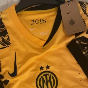Gul Inter Milan x Nike fotbollströja - Helt oanvänd Inter Milan fotbollströja från Nike med svart grafiskt mönster och klubbens emblem på bröstet. Tröjan har korta ärmar, V-ringning och är tillverkad i lätt Dri-FIT material som andas. S passar 170-180