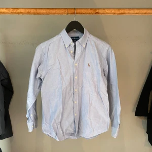 Blå skjorta från Ralph Lauren - Snygg ljusblå skjorta från Ralph Lauren med klassisk krage och knappar hela vägen. Skjortan har lång ärm och den ikoniska broderade loggan på bröstet. Tillverkad i 100% bomull för en skön och fräsch känsla. Perfekt för dig som gillar stilrena och tidlösa plagg. Hör av dig vid minsta fundering!