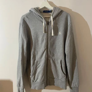 Grå hoodie från Polo Ralph Lauren - Snygg grå hoodie från Polo Ralph Lauren med vit broderad logga på bröstet. Tröjan har huva med vita snören, dragkedja framtill och två stora fickor. Den är väldigt lätt använd och i grymt skick. 