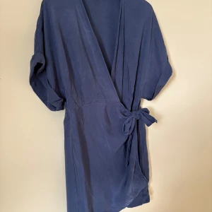 Blå omlottklänning från & Other Stories - Snygg kort omlottklänning från & Other Stories i en djup blå färg. Klänningen har korta ärmar, knyt i midjan och en v-ringad omlottfront. Perfekt för dig som gillar enkel men stilren design med en avslappnad passform.