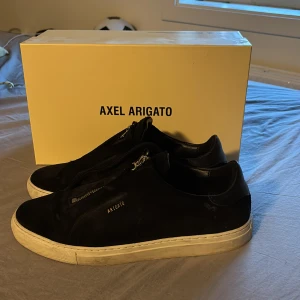 Svarta sneakers Axel Arigato mocka - Svarta sneakers från Axel Arigato i mjuk mocka med vit platt sula. Snygg minimalistisk design med dragkedja framtill istället för klassisk snörning. Diskret Arigato-logga i guld på sidan. Perfekt för dig som gillar stilrena och moderna skor.